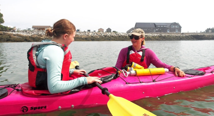 An instructor stands in the water, speaking to a student sitting in a pink kayak. Both people are wearing life jackets. 